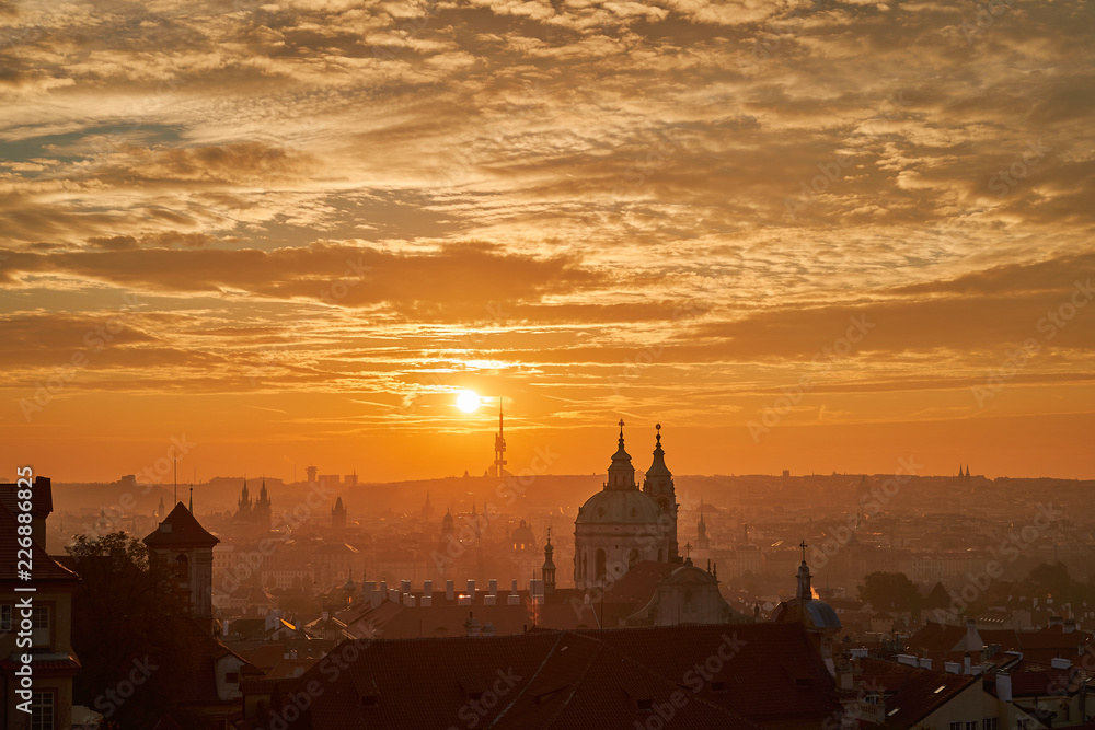 Cityscape of saint Nicolas church in Lesser town, part of downtown or old historic part of Prague in sunrise orange light in morning with sun and golden clouds in sky. 