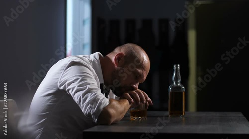 Middle aged alcoholic man thoughtful about alcohol addiction drinking indoors leaning hands on whiskey glass. Depressed lonely adult businessman drinking whiskey trying to forget his problems. Dolly.