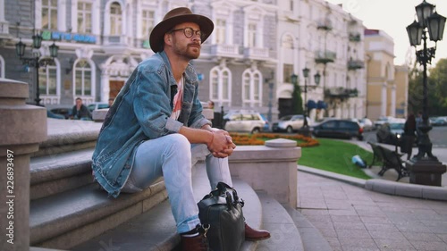 Young stylish handsome redbeard man with bag and in hat sitting on stairs in old city center, slow motion, dolly shot photo