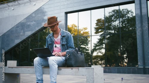 Young bearded hipster man with bag and in hat sits on bench in city center in park using laptop, dolly shot, slow motion photo