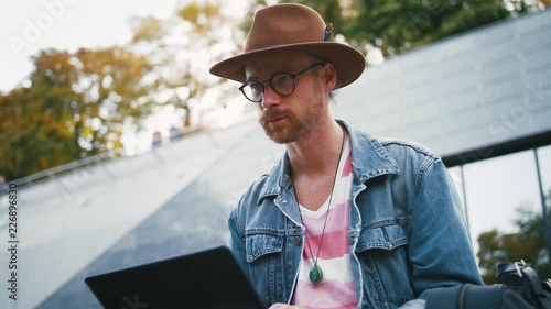 Portrait of young bearded hipster man and in hat sits on bench in city center in park using laptop, slow motion, close up photo