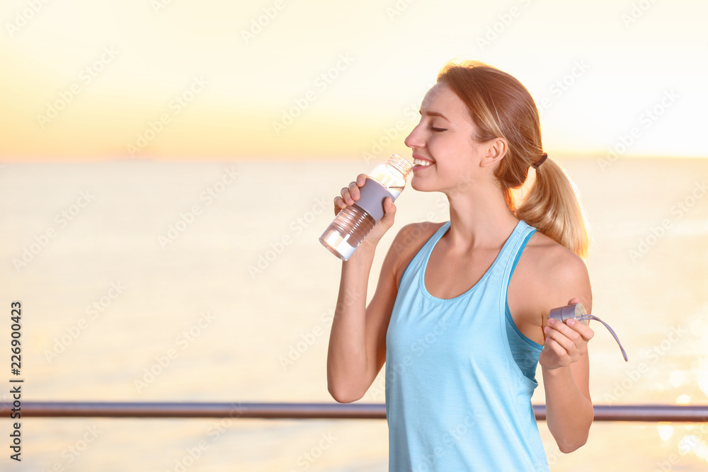 Young woman drinking water from bottle after fitness exercises on pier in morning Young woman drinking water from bottle after fitness exercises on pier in morning