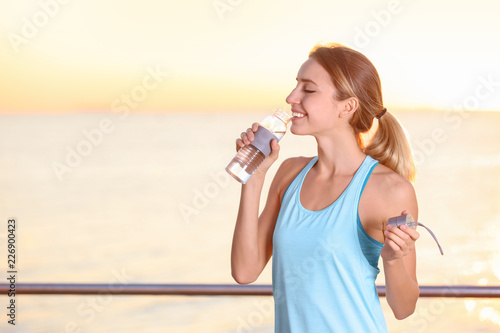Young woman drinking water from bottle after fitness exercises on pier in morning