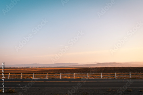 Road and a farm fence with an open farm dry field and mountains background photo