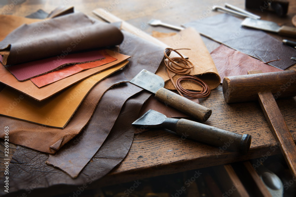 Leather craft or leather working. Selected pieces of beautifully colored or  tanned leather on leather craftman's work desk . Piece of hide and working  tools on a work table. Stock Photo