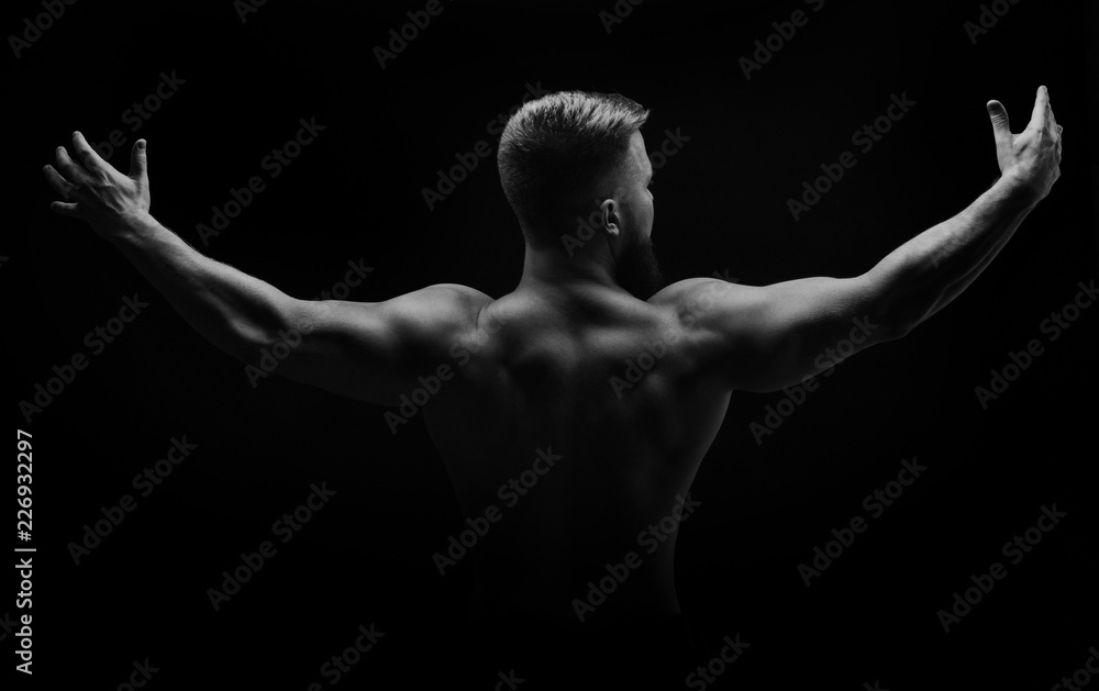 Bodybuilder black and white portrait. Muscular man stands with his back and spread his arms to the side. Broad shoulders. Man unrecognizable