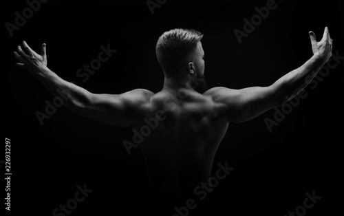 Bodybuilder black and white portrait. Muscular man stands with his back and spread his arms to the side. Broad shoulders. Man unrecognizable