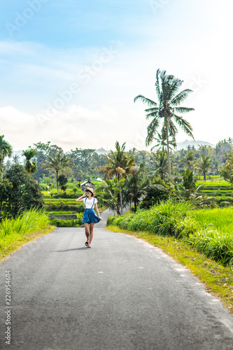 Tropical portrait of young happy woman with straw hat on a road with coconut palms and tropical trees. Bali island.