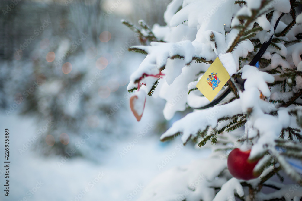 New Jersey state flag. Christmas background outdoor. Christmas tree covered with snow and decorations and New Jersey flag.  New Year / Christmas holiday greeting card.