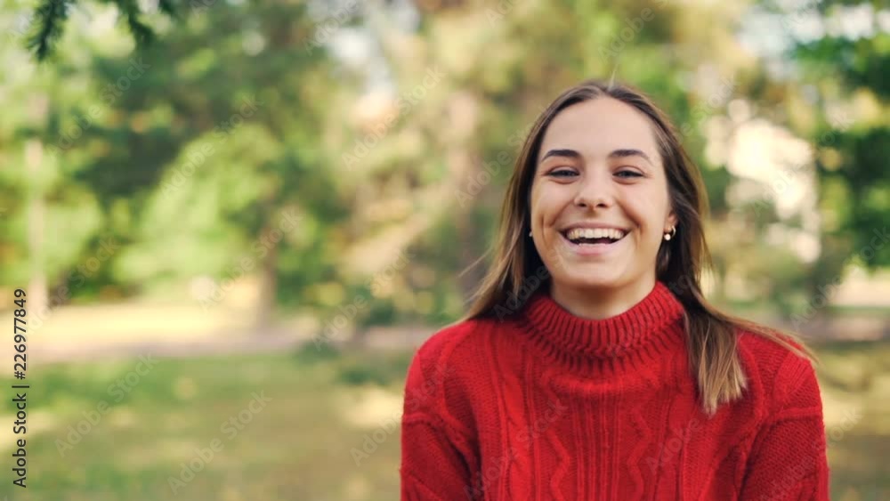 Slow motion portrait of cheerful girl in warm sweater standing in the park with glad smile, laughing and looking at camera. Nature and millennials concept. Slow motion portrait of cheerful girl in warm sweater standing in the park with glad smile, laughing and looking at camera. Nature and millennials concept.