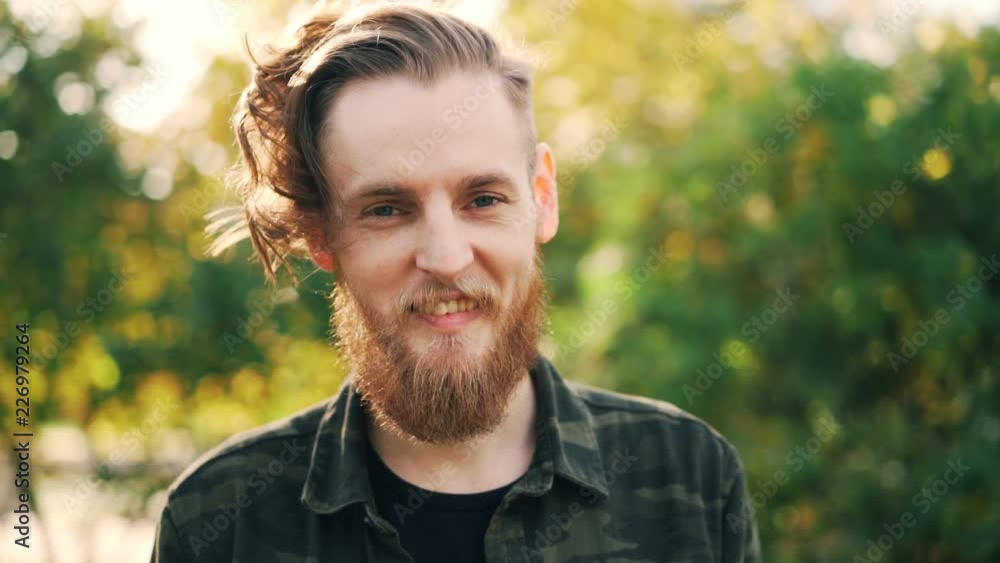Slow motion portrait of carefree guy with fair hair, beard and moustache standing outdoors smiling and looking at camera. People and emotions concept. Slow motion portrait of carefree guy with fair hair, beard and moustache standing outdoors smiling and looking at camera. People and emotions concept.