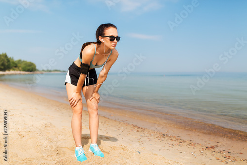 fitness, sport and technology concept - tired female runner with earphones and smartphone in arm band on beach