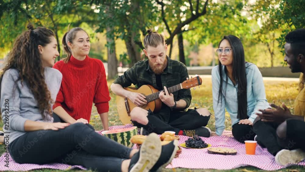 Multiracial group of friends is having fun in park playing the guitar and singing clapping hands enjoying leisure time and warm autumn day. People and music concept. Multiracial group of friends is having fun in park playing the guitar and singing clapping hands enjoying leisure time and warm autumn day. People and music concept.