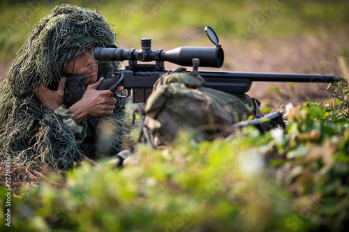 Sniper laying on the grass looking through scope at the target in deep forest.