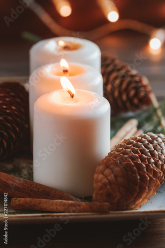 Winter cozy decoration with white burning candles on a wooden festive table and blurrred lights on a background.