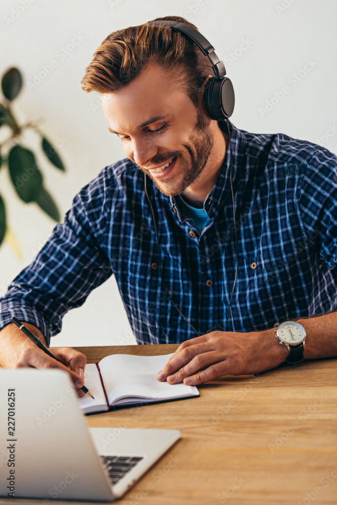 portrait of smiling man in headphones making notes while taking part in webinar at tabletop with laptop in office portrait of smiling man in headphones making notes while taking part in webinar at tabletop with laptop in office