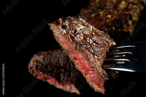 Close up of medium rare beef steak piece on a fork . Spiced with peppercorns and herbs. More fried meat on background. Isolated image with copy space.
