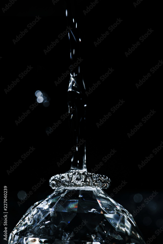 Wedding ring of white gold lying on big diamond and water drop falling from above. Vertical shot with dark black background