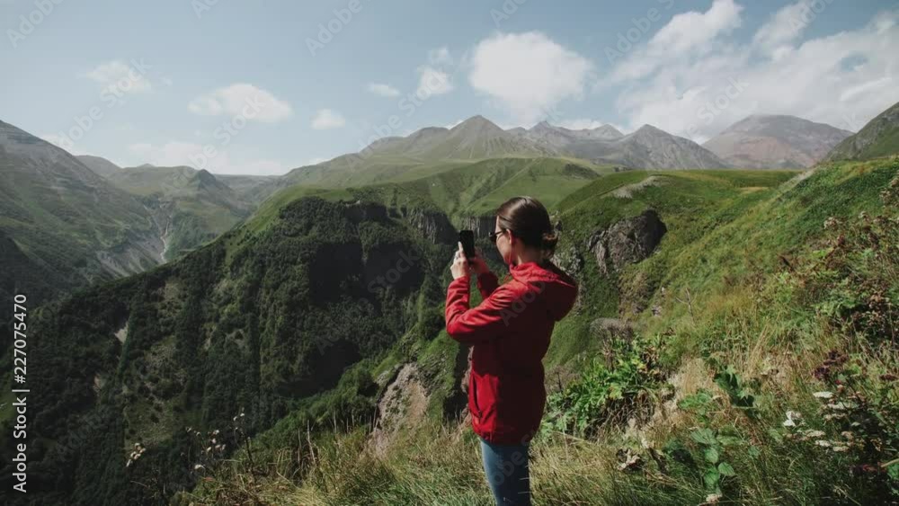 Woman traveler stands on edge of cliff over a mountain gorge. Makes panoramic photo of alpine landscape with hills