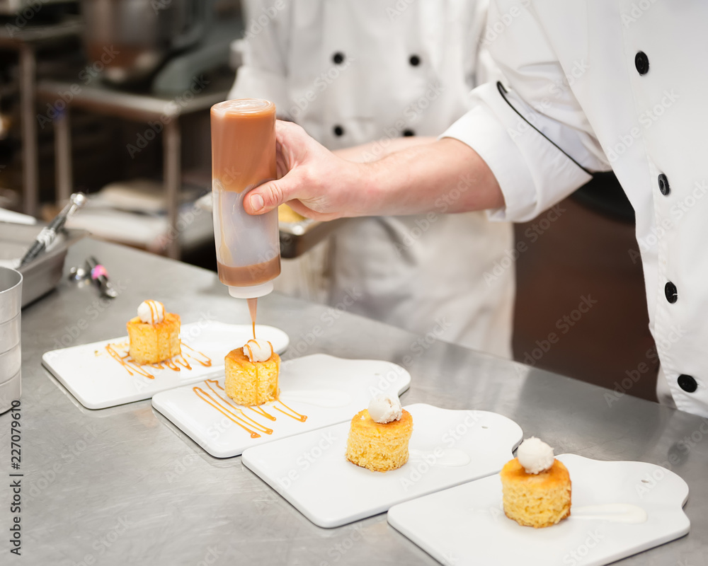 Man in a kitchen drizzling caramel sauce on small cakes topped with cream.