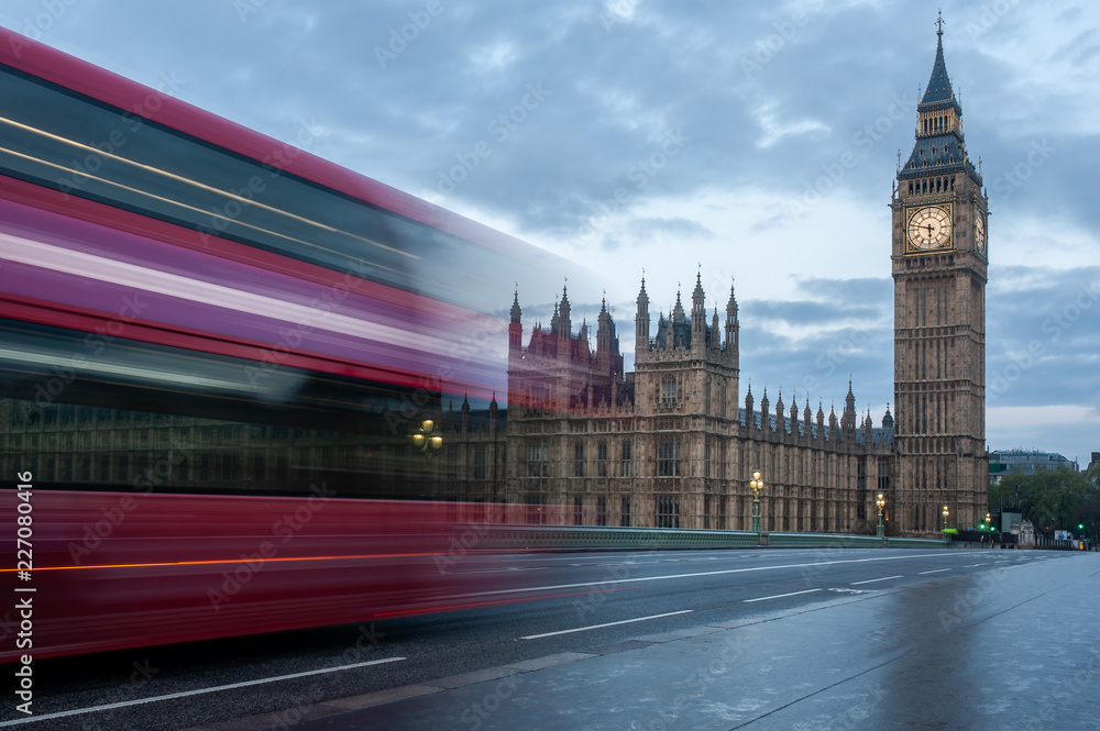 A double-decker bus crosses Westminster Bridge in London at sunrise. No people, nobody. Illuminated Big Ben tower with the clock on the left. Wet pavement. Early morning
