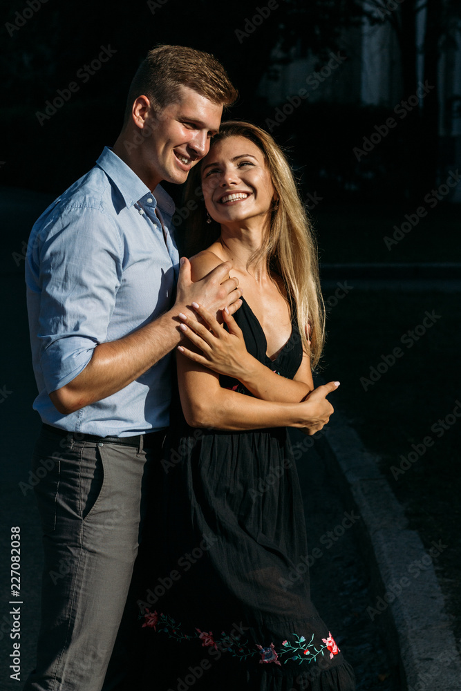 the girl and the guy are smiling holding hands and hugging. the sun shines on their faces. They are on a dark background, shot with hard light.