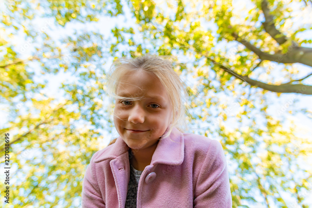 A girl in a pink coat looks into the camera from the top on the background of trees in the park. A girl in a pink coat looks into the camera from the top on the background of trees in the park.
