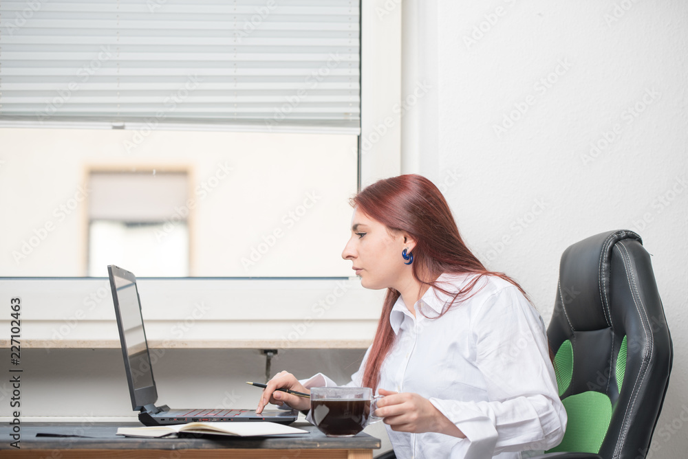 A young woman in a white shirt is sitting at a desk by the window. An open laptop, coffee and notebook are on the desk. The woman is looking for something in the computer, she is concentrated A young woman in a white shirt is sitting at a desk by the window. An open laptop, coffee and notebook are on the desk. The woman is looking for something in the computer, she is concentrated