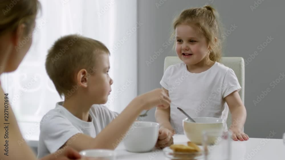 PAN shot of happy little girl sitting at dinner table and talking to mother drinking tea and elder brother eating cereal in morning PAN shot of happy little girl sitting at dinner table and talking to mother drinking tea and elder brother eating cereal in morning