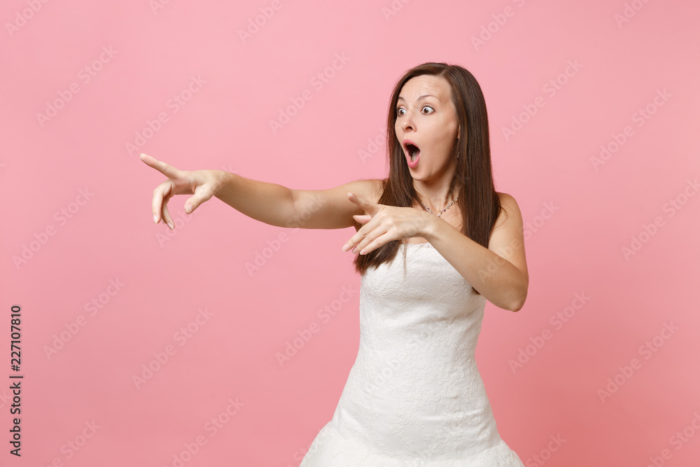 Portrait of shocked bride woman with opened mouth in white wedding dress standing pointing index fingers aside isolated on pink background. Wedding celebration concept. Copy space for advertisement. Portrait of shocked bride woman with opened mouth in white wedding dress standing pointing index fingers aside isolated on pink background. Wedding celebration concept. Copy space for advertisement.