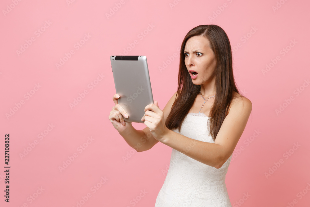 Shocked puzzled bride woman in white wedding dress working on tablet pc computer, making video call isolated on pastel pink background. Wedding to do list. Organization of celebration. Copy space.