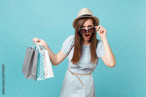 Portrait fashionable attractive happy woman in summer dress, straw hat, sunglasses holding packages bags with purchases after shopping isolated on blue pastel background. Copy space for advertisement.