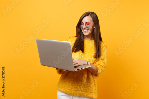 Portrait of smiling young woman in fur sweater and heart eyeglasses working on laptop pc computer isolated on bright yellow background. People sincere emotions, lifestyle concept. Advertising area.