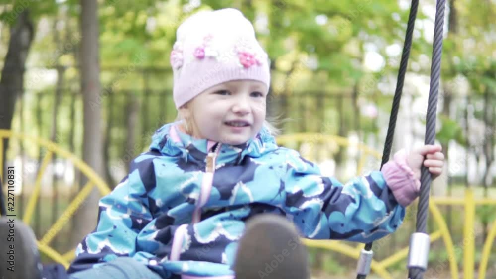 Girl 2 years riding on a swing. On the street is autumn. Baby is warmly dressed. She smiles. the swing goes up and down, forward, back Girl 2 years riding on a swing. On the street is autumn. Baby is warmly dressed. She smiles. the swing goes up and down, forward, back