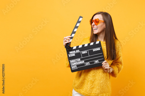 Принти на полотні Laughing young woman in orange heart eyeglasses looking aside and holding classic black film making clapperboard isolated on yellow background