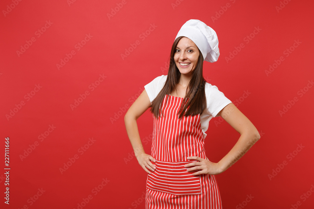 Housewife female chef cook or baker in striped apron, white t-shirt, toque chefs hat isolated on red wall background. Beautiful housekeeper woman standing with arms akimbo. Mock up copy space concept. Housewife female chef cook or baker in striped apron, white t-shirt, toque chefs hat isolated on red wall background. Beautiful housekeeper woman standing with arms akimbo. Mock up copy space concept.