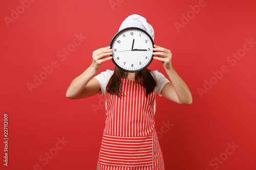 Housewife female chef cook or baker in striped apron white t-shirt toque chefs hat isolated on red wall background. Woman holding in hand in front face round clock hurry up. Mock up copy space concept