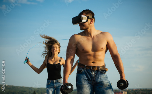 Fototapeta Naklejka Na Ścianę i Meble -  Bodybuilder in vr glasses with dumbbells. Bodybuilder use new technology for training. Bodybuilder build muscle while woman skipping on blue sky. Bodybuilder with fit torso. In touch with tomorrow