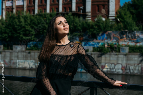 beautiful girl, brunette, in the summer by the river in the city, in a black dress. standing alone by the river, on the bridge, the sun is shining in the face, closed her eyes, rest and tourism.