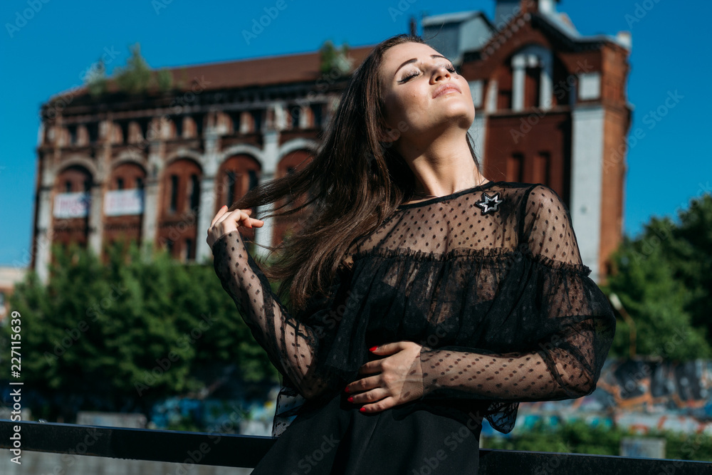 beautiful girl, brunette, in the summer by the river in the city, in a black dress. standing alone by the river, on the bridge, the sun is shining in the face, closed her eyes, rest and tourism.