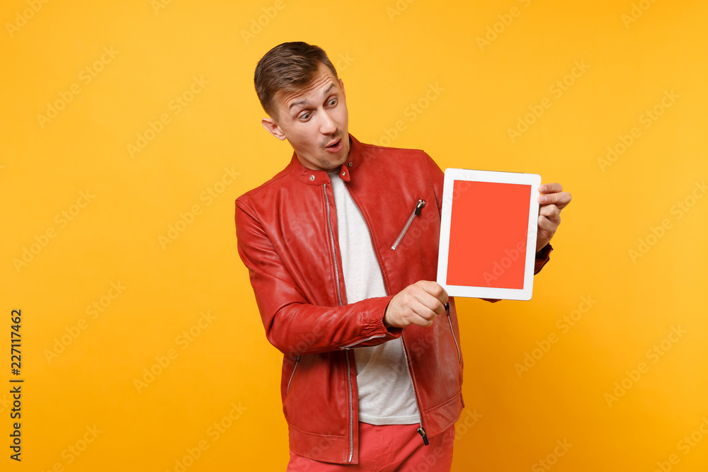Portrait vogue fun young man in red leather jacket, t-shirt hold tablet pc pad with blank empty screen isolated on bright yellow background. People sincere emotions lifestyle concept. Advertising area Portrait vogue fun young man in red leather jacket, t-shirt hold tablet pc pad with blank empty screen isolated on bright yellow background. People sincere emotions lifestyle concept. Advertising area