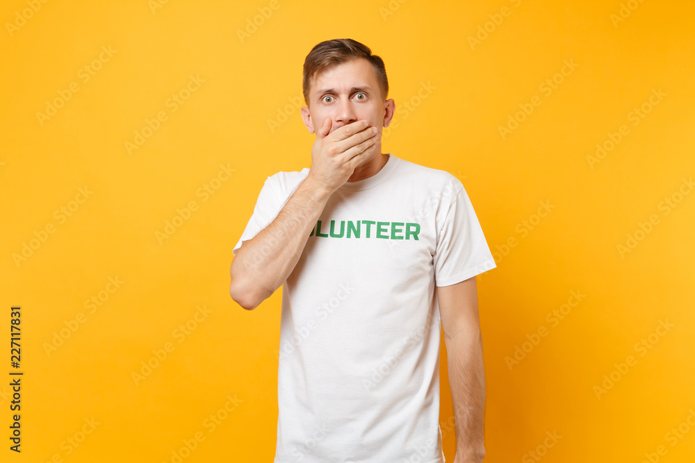 Portrait of surprised shocked fun young man in white t-shirt with written inscription green title volunteer isolated on yellow background. Voluntary free assistance help, charity grace work concept. Portrait of surprised shocked fun young man in white t-shirt with written inscription green title volunteer isolated on yellow background. Voluntary free assistance help, charity grace work concept.