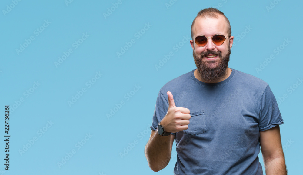 Young caucasian hipster man wearing sunglasses over isolated background doing happy thumbs up gesture with hand. Approving expression looking at the camera with showing success. Young caucasian hipster man wearing sunglasses over isolated background doing happy thumbs up gesture with hand. Approving expression looking at the camera with showing success.