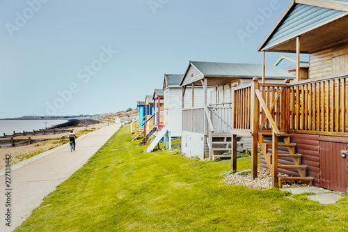 Quaint wooden beach huts in a row along a grass verge in front of the promenade that a man is cycling along, by the beach and sea in Tankerton, Whitstable, Kent, UK photo