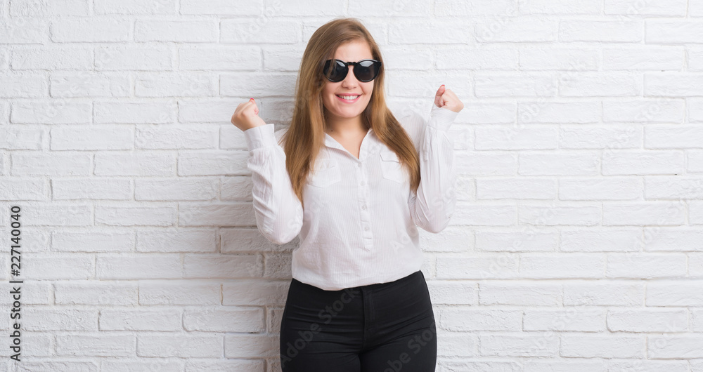 Young adult business woman standing over white brick wall wearing sunglasses screaming proud and celebrating victory and success very excited, cheering emotion Young adult business woman standing over white brick wall wearing sunglasses screaming proud and celebrating victory and success very excited, cheering emotion