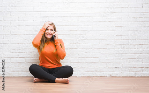 Young adult woman sitting on the floor over white brick wall calling using smartphone stressed with hand on head, shocked with shame and surprise face, angry and frustrated. Fear and upset