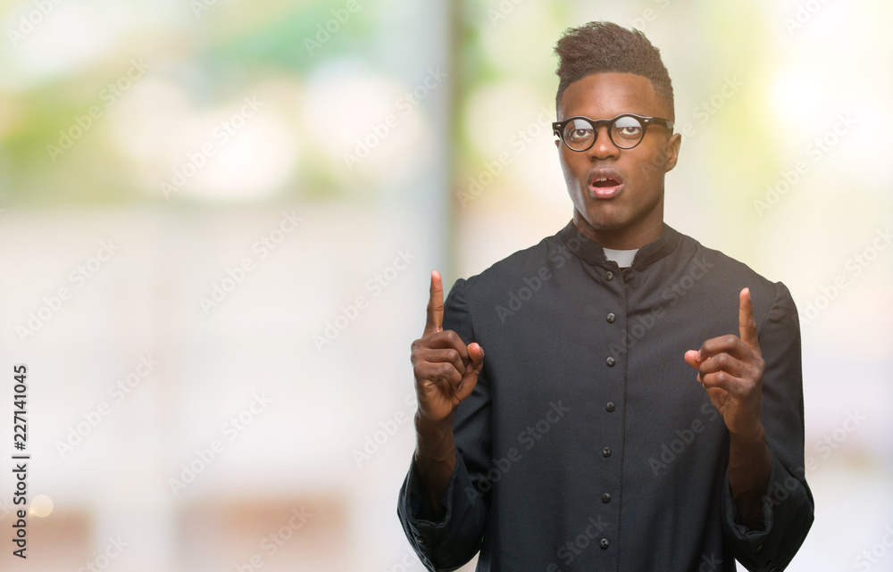 Young african american priest man over isolated background amazed and surprised looking up and pointing with fingers and raised arms. Young african american priest man over isolated background amazed and surprised looking up and pointing with fingers and raised arms.