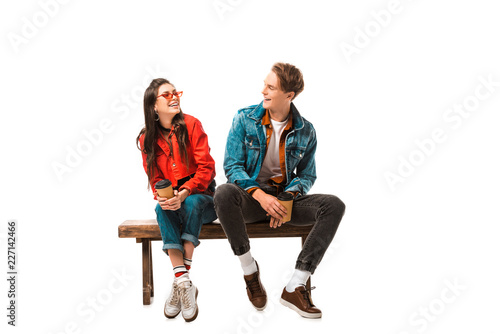 Fotobehang laughing hipster couple with disposable cups of coffee sitting on bench isolated