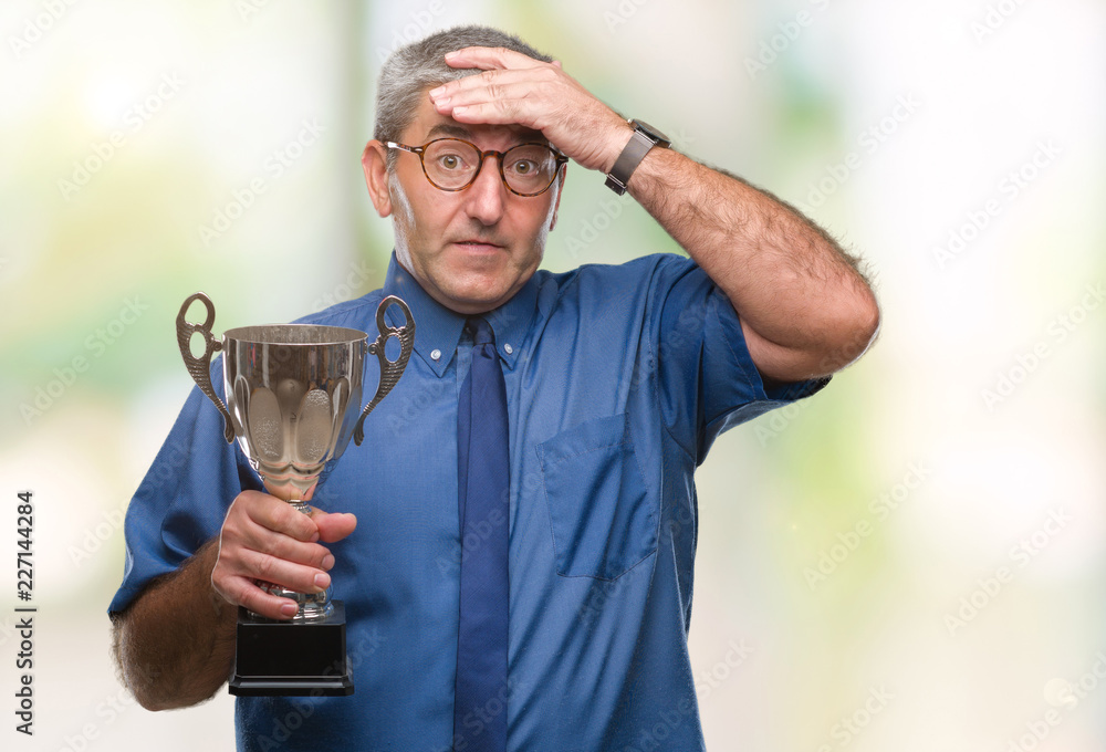 Handsome senior successful man holding trophy over isolated background stressed with hand on head, shocked with shame and surprise face, angry and frustrated. Fear and upset for mistake. Handsome senior successful man holding trophy over isolated background stressed with hand on head, shocked with shame and surprise face, angry and frustrated. Fear and upset for mistake.