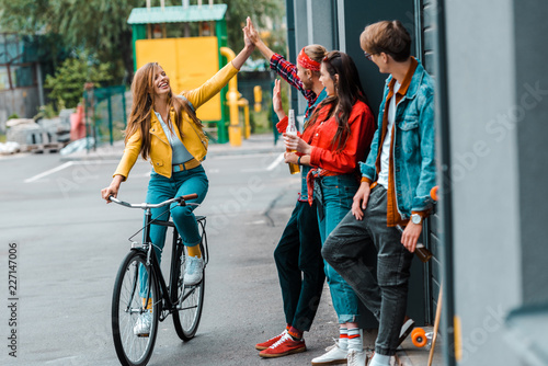 happy stylish girl riding bike and giving highfive to friends on street